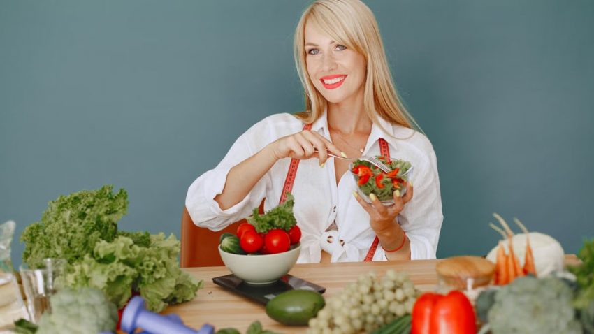 Blonde woman enjoying a fresh salad surrounded by vegetables, promoting healthy eating.